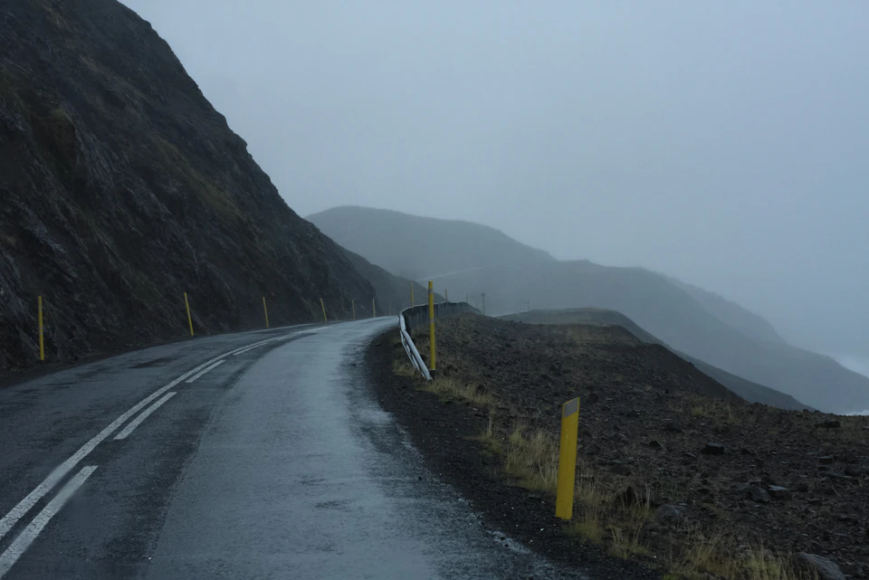 The image depicts a winding road cutting through a mountainous landscape, with a foggy and overcast sky above. The road is flanked by lush green grass on both sides, leading to a hill covered in rocks and vegetation at the bottom. On either side of the road are yellow poles marking the boundary between the two lanes.