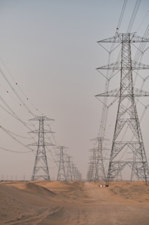 A series of large electricity pylons extends into the distance over a sandy desert landscape. The pylons are evenly spaced and connected by numerous power lines. The sky above is clear and pale, contributing to the sense of openness and expanse.