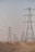 A series of large electricity pylons extends into the distance over a sandy desert landscape. The pylons are evenly spaced and connected by numerous power lines. The sky above is clear and pale, contributing to the sense of openness and expanse.