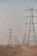 A series of large electricity pylons extends into the distance over a sandy desert landscape. The pylons are evenly spaced and connected by numerous power lines. The sky above is clear and pale, contributing to the sense of openness and expanse.
