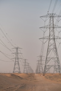 A series of large electricity pylons extends into the distance over a sandy desert landscape. The pylons are evenly spaced and connected by numerous power lines. The sky above is clear and pale, contributing to the sense of openness and expanse.