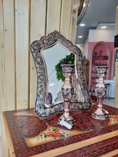 An ornately designed mirror with intricate floral patterns stands on a carved wooden surface, accompanied by two elegant candlesticks. The background features vibrant pink walls and various decorative items on shelves. The wooden surface has detailed carvings and painted floral motifs.