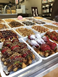 A display of various types of waffles on a counter, each topped with different ingredients such as strawberries, powdered sugar, chocolate drizzle, and other toppings. The counter has multiple white trays filled with these waffles, and a sign indicating a price of 2 euros is visible in the background. The setting appears to be a bakery or a food stall.
