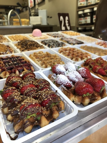 A display of various types of waffles on a counter, each topped with different ingredients such as strawberries, powdered sugar, chocolate drizzle, and other toppings. The counter has multiple white trays filled with these waffles, and a sign indicating a price of 2 euros is visible in the background. The setting appears to be a bakery or a food stall.