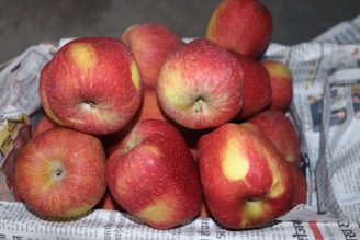 A pile of red apples with yellow patches arranged in a basket lined with newspapers, displaying a fresh and natural appearance.
