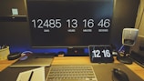A desk setup featuring a large monitor displaying a countdown of days, hours, minutes, and seconds. Below the monitor, there is a laptop, a wireless keyboard, and a mouse. A small tablet also shows the time. On the right, a retro-style microphone is placed. The workspace is organized with a notebook, a pen, and a pot with pens.