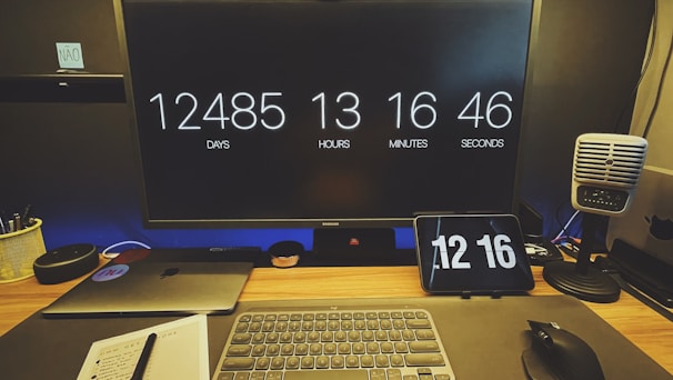 A desk setup featuring a large monitor displaying a countdown of days, hours, minutes, and seconds. Below the monitor, there is a laptop, a wireless keyboard, and a mouse. A small tablet also shows the time. On the right, a retro-style microphone is placed. The workspace is organized with a notebook, a pen, and a pot with pens.