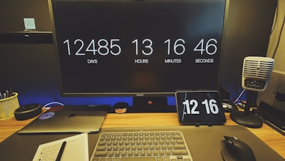 A desk setup featuring a large monitor displaying a countdown of days, hours, minutes, and seconds. Below the monitor, there is a laptop, a wireless keyboard, and a mouse. A small tablet also shows the time. On the right, a retro-style microphone is placed. The workspace is organized with a notebook, a pen, and a pot with pens.