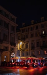 A nighttime scene of a lively outdoor cafe with people seated under large umbrellas and warm lighting, set against the backdrop of tall, ornate buildings.