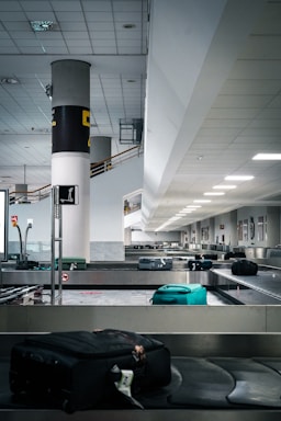 An airport baggage claim area features a conveyor belt with several suitcases, mainly in dark and turquoise colors. The hall is spacious and modern, with a high ceiling and fluorescent lighting. A column with signage stands to the left, and the area appears quiet and empty.