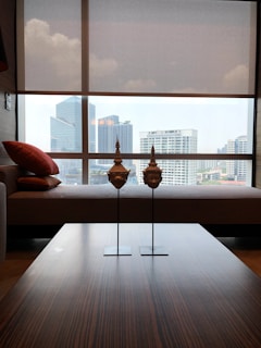 A modern interior with a large window overlooking a cityscape featuring several high-rise buildings. In the foreground, there is a wooden table with two decorative sculptures resembling traditional masks. To the left, a sofa adorned with red cushions is visible.