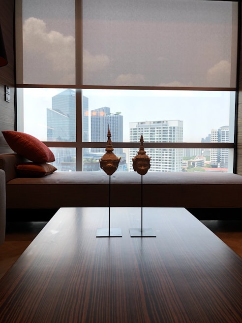A modern interior with a large window overlooking a cityscape featuring several high-rise buildings. In the foreground, there is a wooden table with two decorative sculptures resembling traditional masks. To the left, a sofa adorned with red cushions is visible.