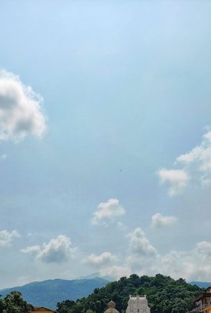 A lush green hill adorned with an ornate temple structure rises against a backdrop of a clear sky dotted with fluffy white clouds. The scene conveys a sense of serenity and natural beauty.