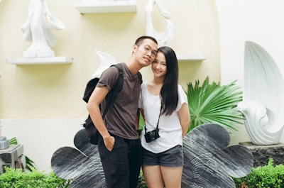 A smiling couple stands close together in an outdoor setting, surrounded by large sculptural elements and greenery. The man wears glasses, a dark shirt, and a backpack, while the woman has long dark hair, a white shirt, and a camera hanging around her neck.