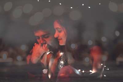 A tender and intimate moment is captured between a couple surrounded by a soft, dreamy bokeh effect. The man appears contemplative, resting his chin on his hand, while the woman looks at him with a gentle smile. Warm lighting creates a cozy and romantic atmosphere.