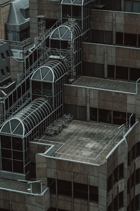 A rooftop section of a modern building featuring geometric architectural designs with a combination of glass and metal structures. Several empty chairs are arranged on a tiled terrace, surrounded by a railing. The overall structure includes multiple levels with prominent glass domes and intricate metal frameworks.