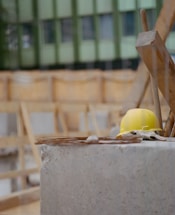 A yellow hard hat rests on a concrete surface at a construction site. The background shows unfinished wooden structures and a green building facade.