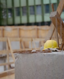A yellow hard hat rests on a concrete surface at a construction site. The background shows unfinished wooden structures and a green building facade.