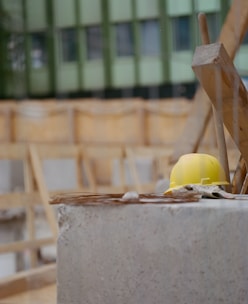 A yellow hard hat rests on a concrete surface at a construction site. The background shows unfinished wooden structures and a green building facade.