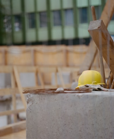 A yellow hard hat rests on a concrete surface at a construction site. The background shows unfinished wooden structures and a green building facade.