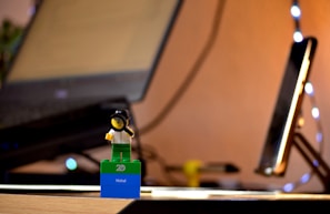 A small LEGO figurine holding a magnifying glass stands on a blue and green base. The base has the word 'Nishal' written on it. In the blurred background, there is a laptop and a tablet with fairy lights adding a warm glow to the scene. The setting appears to be a desk or workspace.