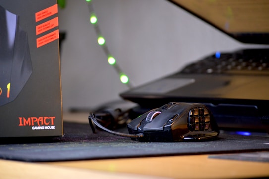 A gaming mouse with multiple buttons is placed on a mouse pad next to a laptop. The box for the mouse, labeled 'IMPACT GAMING MOUSE,' is visible on the left side. In the background, there are faint lights creating an ambient atmosphere.
