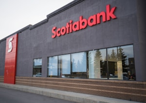 The facade of a building with a sign displaying 'Scotiabank' in large red letters. There are reflective glass windows and a mix of smooth grey and brick wall surfaces. The image includes a portion of the sidewalk and some trees reflected on the glass.