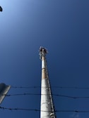 A tall communication tower is positioned against a clear blue sky. The perspective is upward-facing, emphasizing the height of the structure. Barbed wire is visible in the foreground, adding a sense of restriction or security.