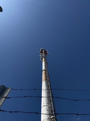 A tall communication tower is positioned against a clear blue sky. The perspective is upward-facing, emphasizing the height of the structure. Barbed wire is visible in the foreground, adding a sense of restriction or security.