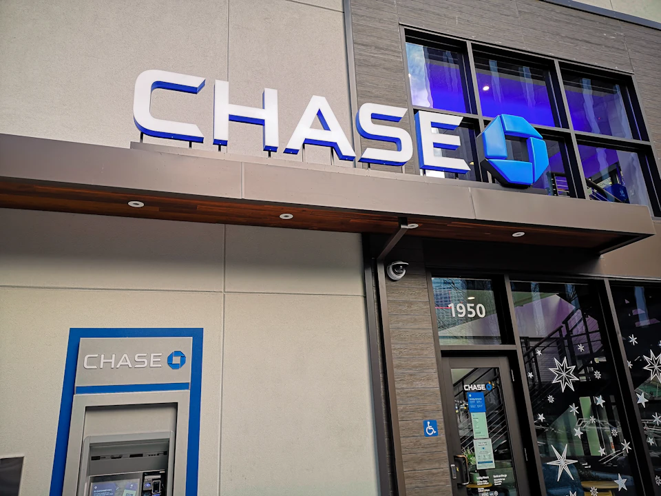 The image shows a Chase Bank building with a large blue sign displaying the Chase logo on its facade. The bank's name is prominently displayed in white letters above the sign, and the building features a gray exterior with a brown roof. A glass window can be seen on one side of the building, reflecting the surroundings.