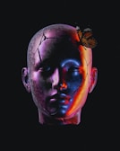A sculpture-like human head with visible cracks and surreal, colorful lighting. A butterfly is perched gracefully on the head, offering a contrast between fragility and resilience.