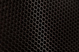A close-up view of a metal mesh with a hexagonal pattern creating a honeycomb-like structure. The mesh is in a dark color against a black background, showcasing its intricate geometric design.