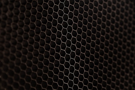 A close-up view of a metal mesh with a hexagonal pattern creating a honeycomb-like structure. The mesh is in a dark color against a black background, showcasing its intricate geometric design.