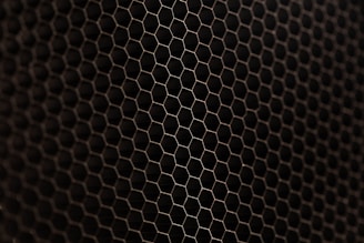 A close-up view of a metal mesh with a hexagonal pattern creating a honeycomb-like structure. The mesh is in a dark color against a black background, showcasing its intricate geometric design.