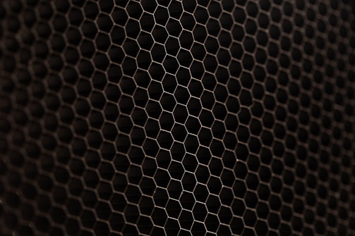 A close-up view of a metal mesh with a hexagonal pattern creating a honeycomb-like structure. The mesh is in a dark color against a black background, showcasing its intricate geometric design.