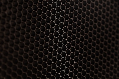 A close-up view of a metal mesh with a hexagonal pattern creating a honeycomb-like structure. The mesh is in a dark color against a black background, showcasing its intricate geometric design.