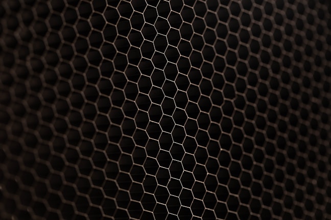 A close-up view of a metal mesh with a hexagonal pattern creating a honeycomb-like structure. The mesh is in a dark color against a black background, showcasing its intricate geometric design.