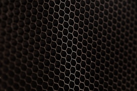 A close-up view of a metal mesh with a hexagonal pattern creating a honeycomb-like structure. The mesh is in a dark color against a black background, showcasing its intricate geometric design.