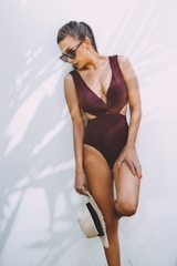 A woman wearing a maroon one-piece swimsuit and sunglasses is standing against a plain white wall. She is holding a straw hat with a black ribbon in one hand and has her hair tied back in a ponytail.