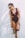 A woman wearing a maroon one-piece swimsuit and sunglasses is standing against a plain white wall. She is holding a straw hat with a black ribbon in one hand and has her hair tied back in a ponytail.