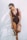 A woman wearing a maroon one-piece swimsuit and sunglasses is standing against a plain white wall. She is holding a straw hat with a black ribbon in one hand and has her hair tied back in a ponytail.
