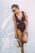 A woman wearing a maroon one-piece swimsuit and sunglasses is standing against a plain white wall. She is holding a straw hat with a black ribbon in one hand and has her hair tied back in a ponytail.