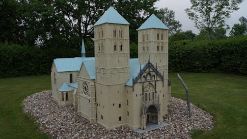 A detailed scale model of a cathedral featuring two tall towers with blue roofs, intricate architectural details, and positioned on a gravel base surrounded by lush green grass. The model is placed outdoors among trees and bushes, suggesting a garden or park setting.