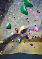 A person is climbing an indoor rock-climbing wall with various colored holds. The wall features a mix of green, pink, yellow, and purple climbing holds randomly distributed across a textured surface. The person appears focused and is wearing climbing attire.