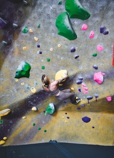 A person is climbing an indoor rock-climbing wall with various colored holds. The wall features a mix of green, pink, yellow, and purple climbing holds randomly distributed across a textured surface. The person appears focused and is wearing climbing attire.