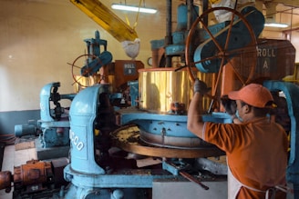 A person wearing an orange cap and shirt operates industrial machinery with large gears and wheels in a factory setting. The equipment is predominantly blue with copper elements, and bags are suspended overhead. The environment appears to be industrial, with a focus on mechanical processing.