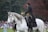 A person dressed in an ornate, dark outfit rides a white horse in a grassy outdoor area. The horse is elegantly poised, with its mane flowing. In the background, there is a blurred crowd of people, suggesting a public event or performance. Trees can be seen in the distance, adding a natural setting to the scene.
