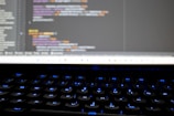 A close-up view of a computer screen displaying lines of colorful programming code. The keyboard in the foreground is slightly blurred, with keys lit by blue backlighting. The code on the screen includes syntax highlighting with various colors used to differentiate between components.