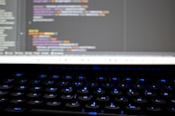 A close-up view of a computer screen displaying lines of colorful programming code. The keyboard in the foreground is slightly blurred, with keys lit by blue backlighting. The code on the screen includes syntax highlighting with various colors used to differentiate between components.