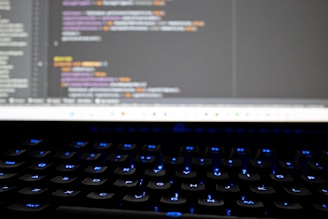 A close-up view of a computer screen displaying lines of colorful programming code. The keyboard in the foreground is slightly blurred, with keys lit by blue backlighting. The code on the screen includes syntax highlighting with various colors used to differentiate between components.